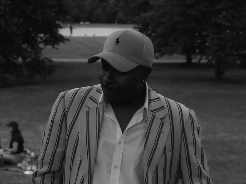 A man in a striped shirt and cap standing in a park, black and white