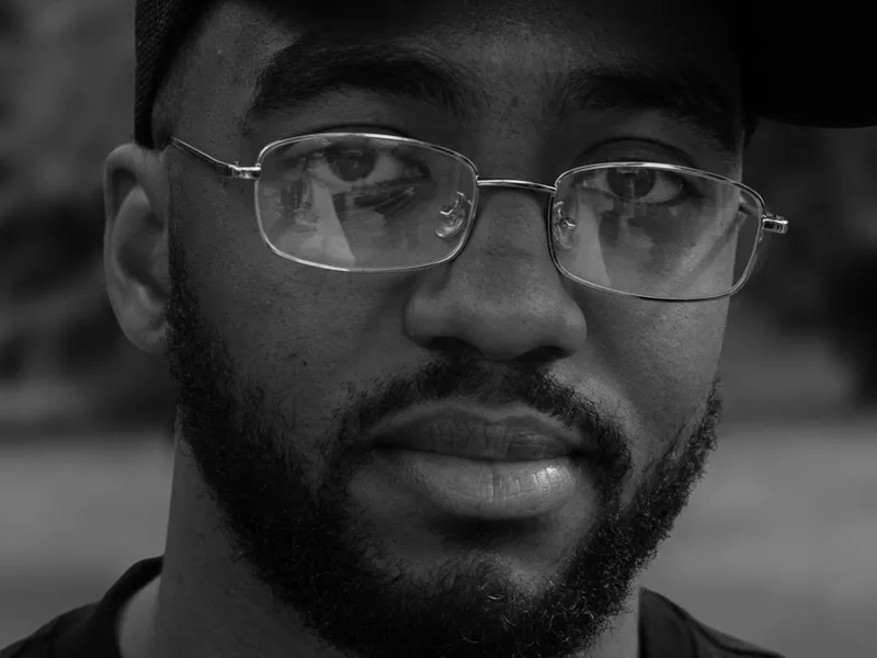 A close black and white portrait of a man in glasses and a slogan cap