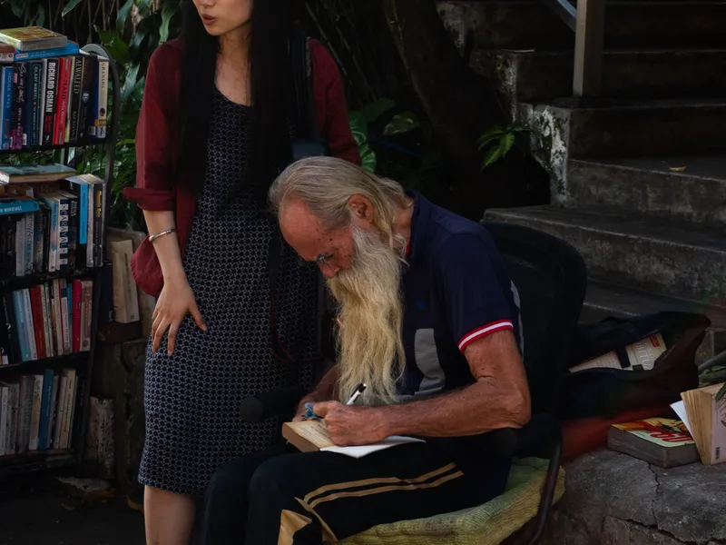 A woman standing beside an older man writing at a roadside bookstall
